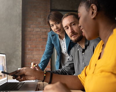 A group of people in a casual office setting, collaborating and discussing something on a laptop screen. The focus is on teamwork and collaboration, with a diverse group of individuals contributing to the discussion.