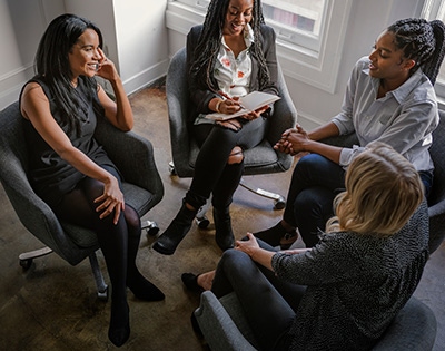 A group of four women sitting in a circle on chairs, engaged in a discussion. They are in a well-lit room with large windows. The women are of diverse ethnic backgrounds, and one of them is taking notes in a notebook.